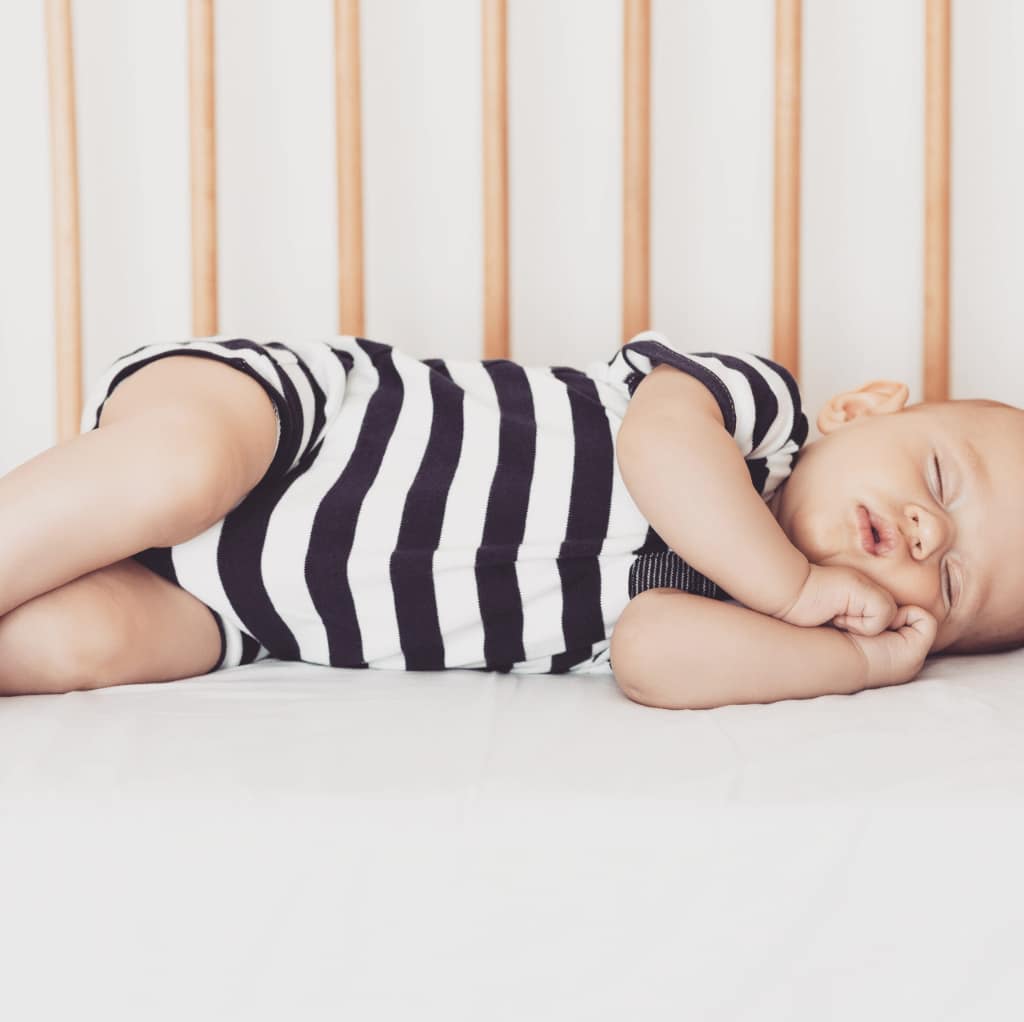Baby sleeping on it's side in a wooden cot wearing a black and white striped baby grow with hands cupped together near face